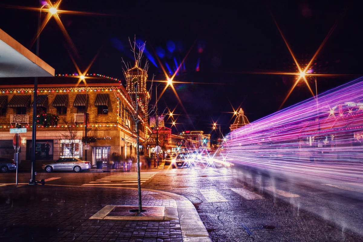 Next Guest KC management fees, Spanish-tiled shopping district lit up at night with traffic trails