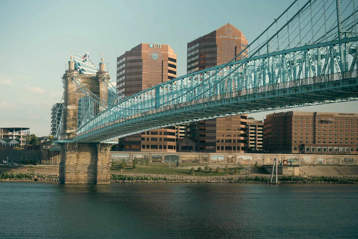 Stay Cincinnati management fees, Teal suspension bridge spanning the river below downtown buildings