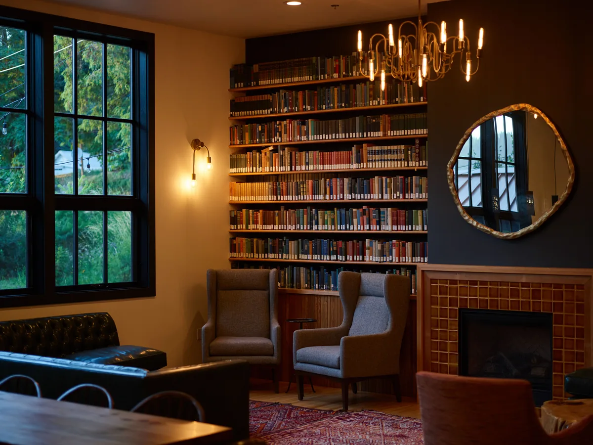 Classic interior design of spacious living room in crural cottage with big window and comfy armchairs near big bookshelves and round vintage mirror above cozy fireplace against classy chandelier